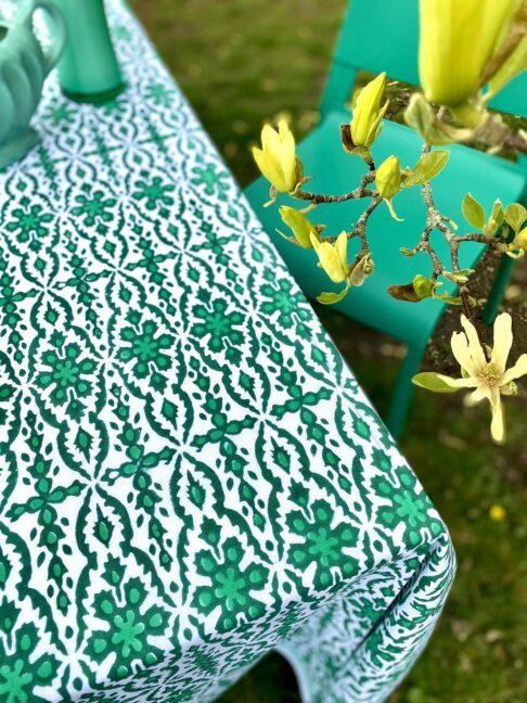 A green patterned tablecloth with an Indian block print design styled with green vases and a chair in an outdoor setting.