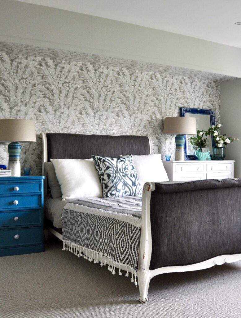 A grey-upholstered, French sleigh bed against 'Florencecourt' wallpaper by Cole & Son in a calm, bedroom with teal blue accents.