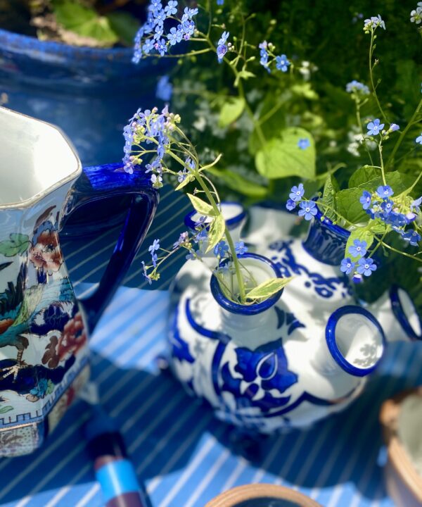 A Dutch tulip vase filled with wild forget-me-nots on an outdoor table with a blue and white striped tablecloth and assorted vintage china jugs.