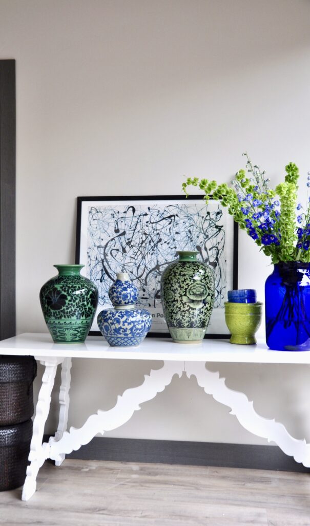 A group of blue and green pots and vases on a white refectory table with a Jackson Pollack print behind.