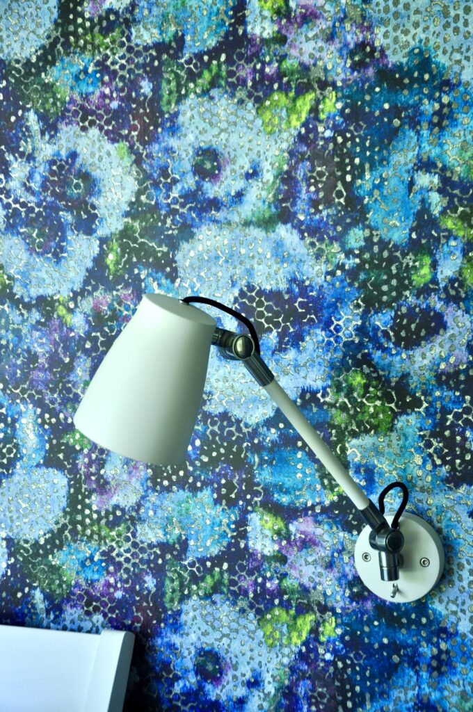 A simple, white, adjustable wall lamp against a bold, modern , floral wallpaper in blue and green by Designers Guild.