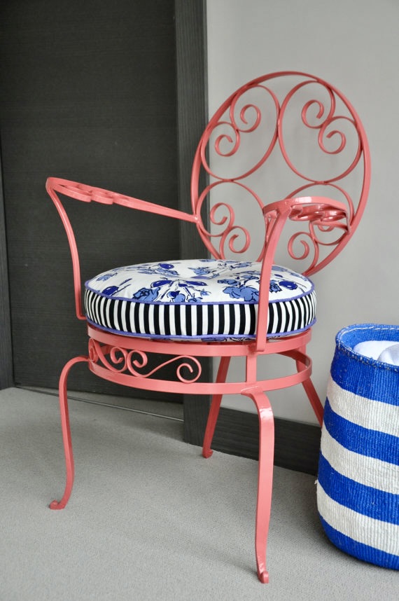 A French, antique wrought iron chair re-sprayed in coral pink with a circular floral and stripe cushion and a blue and white striped basket in the foreground.