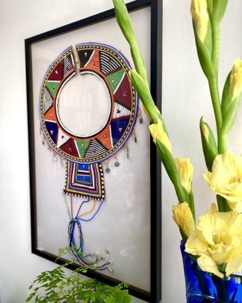 A framed Maasai beadwork collar necklace with yellow gladioli in a vase in the foreground.