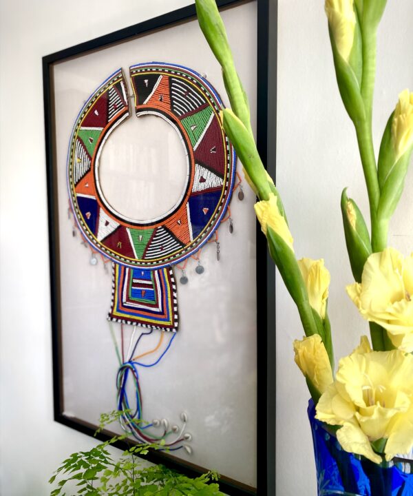 A framed Maasai beadwork collar necklace with yellow gladioli in a vase in the foreground.