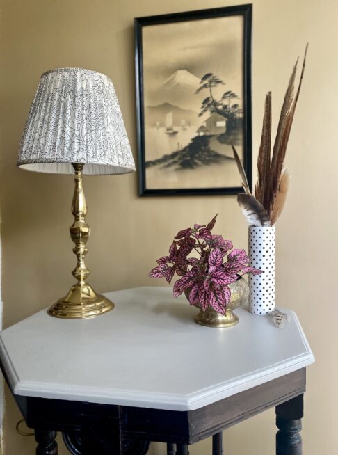 A gathered table lampshade on a brass base with decorative accessories on an octagonal side table with a top painted in pale grey.