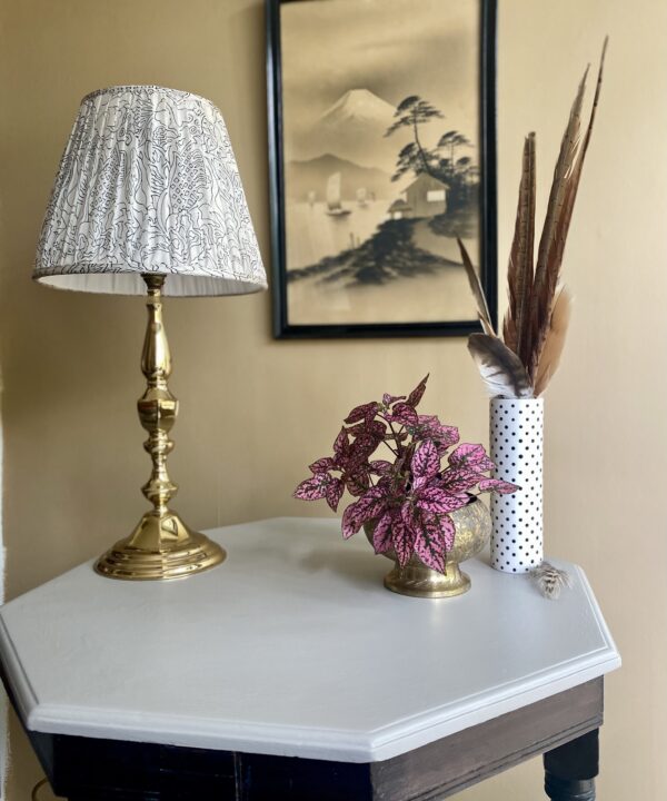 A gathered table lampshade on a brass base with decorative accessories on an octagonal side table with a top painted in pale grey.