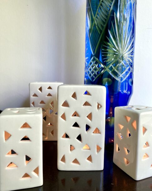 A collection of white ceramic, pierced tea light holders in front of a blue, cut glass vase.