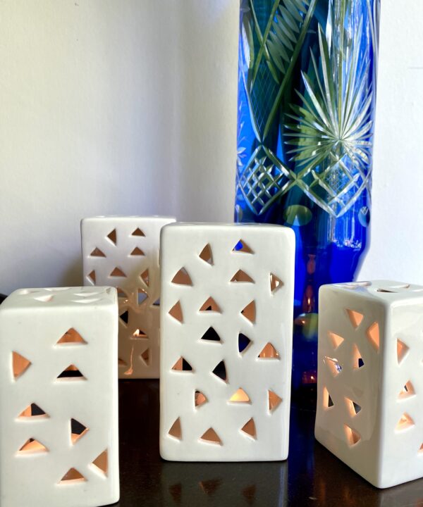 A collection of white ceramic, pierced tea light holders in front of a blue, cut glass vase.