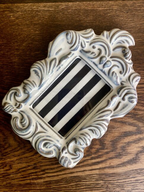 A decorative white ceramic photo frame from Yarnnakarn ceramics studio Bangkok, with a scrolling, acanthus leaf design on a wooden table.