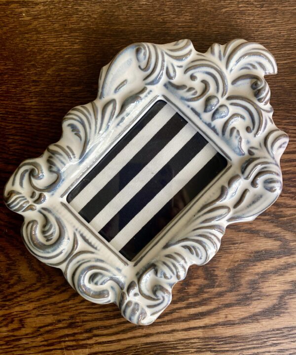 A decorative white ceramic photo frame from Yarnnakarn ceramics studio Bangkok, with a scrolling, acanthus leaf design on a wooden table.