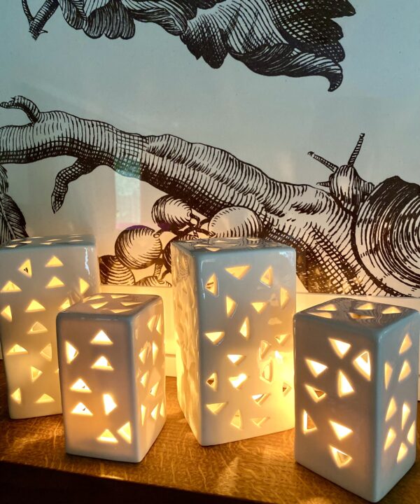 A group pf glowing, pierced, ceramic tea light holders on a shelf in front of a large scale print of of a snail on a tree.