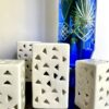 A group of white ceramic, pierced tea light holders in front of a blue, cut glass vase.