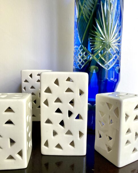 A group of white ceramic, pierced tea light holders in front of a blue, cut glass vase.