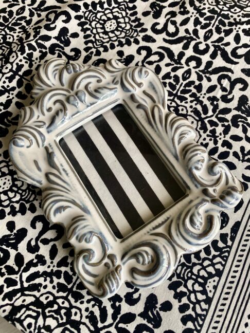 A white china photograph frame with decorative, handmade acanthus leaf scrolls against a black and white patterned table cloth.