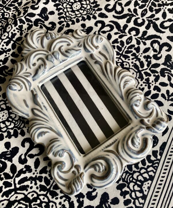 A white china photograph frame with decorative, handmade acanthus leaf scrolls against a black and white patterned table cloth.