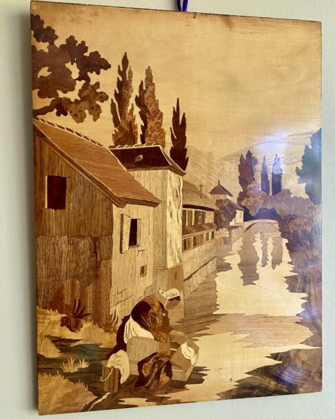 A vintage, wooden inlay wall art panel depicting a canalside scene with a woman washing laundry.