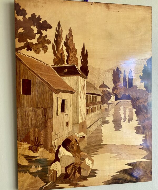 A vintage, wooden inlay wall art panel depicting a canalside scene with a woman washing laundry.