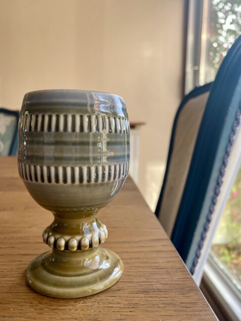 A small, handmade Irish craft pottery bud vase or goblet by Wade, sat on a dining table with a chair in the background.