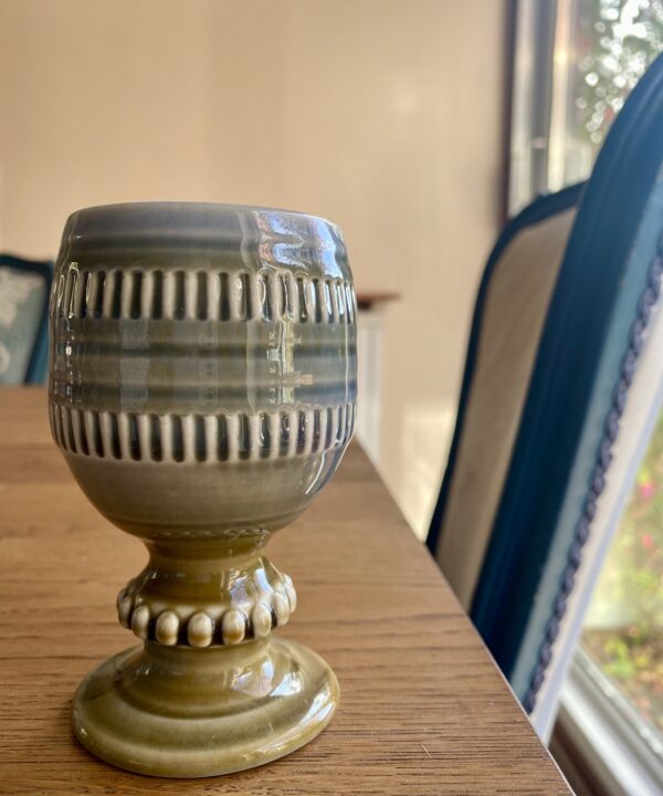 A small, handmade Irish craft pottery bud vase or goblet by Wade, sat on a dining table with a chair in the background.