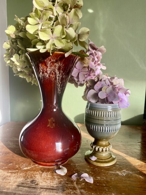 A small, brown, retro vase handmade at the Kernewek pottery in Cornwall likely during the 1970s. Styled here with hydrangeas on a wooden table.