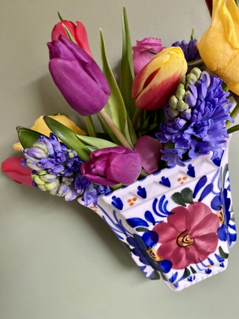 Hyacinths, roses and variegated tulips fill a vintage Portuguese painted vase, hanging on a wall.